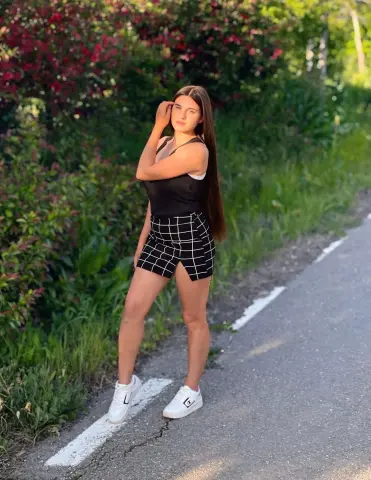 Young woman in a black tank top and plaid mini skirt posing on a sunny garden path.