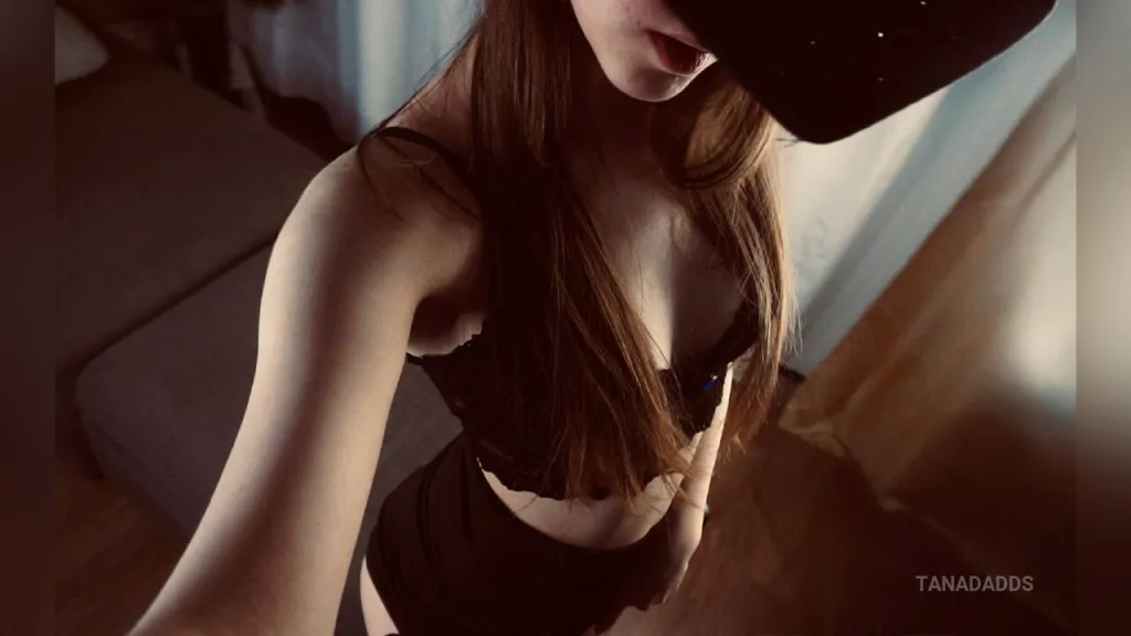 TanaDadds poses with her eyes closed, holding her breasts with her hands in a dimly lit room.