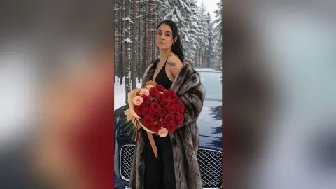 Elegant woman in a black dress and fur coat holding a large red rose bouquet in a snowy winter forest.