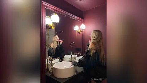 Blonde woman in a black leather jacket applying lipstick in a moody, modern bathroom with warm lighting.