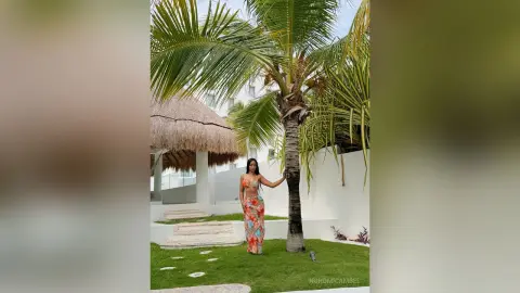 NohomiCambel stands next to a palm tree in a long, floral print sundress against a white wall.