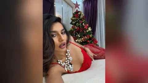 Glamorous woman in a red lace outfit and chunky white necklace relaxing near a decorated Christmas tree.