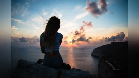 AnaMathers sits on a rocky cliffside overlooking the ocean at sunset, her long blonde hair blowing in the wind.