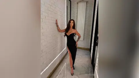 ReynaGomes poses in a tight black strapless dress with a high slit, leaning against a white brick wall in a bright room.
