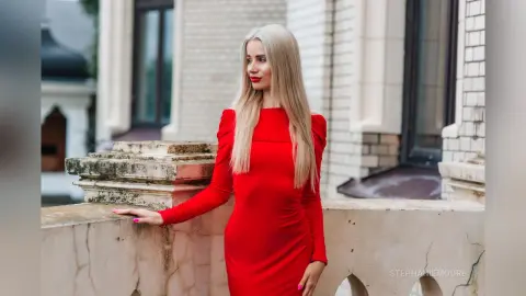 Elegant blonde woman in a red long-sleeve dress posing by a stone balcony of a classic building.