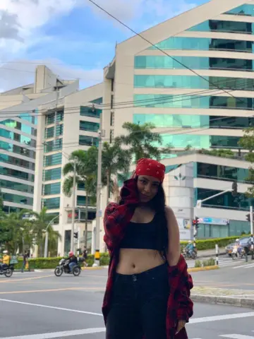 Young woman in a red plaid shirt and bandana posing on a sunny city street near modern glass buildings.