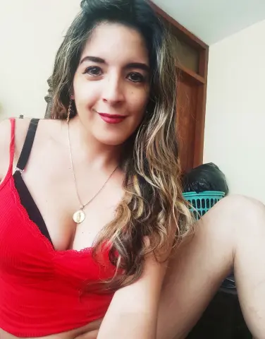 Smiling woman in a red tank top and gold necklace relaxing indoors with long wavy hair.