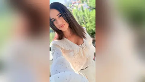 BellaLeen poses outdoors in a white polka-dot blouse that shows off her cleavage, looking at the camera with long brown hair.