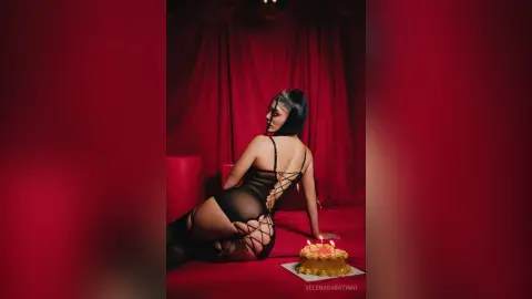 Elegant woman in black lace bodysuit sitting by a birthday cake against red velvet curtains in moody lighting.