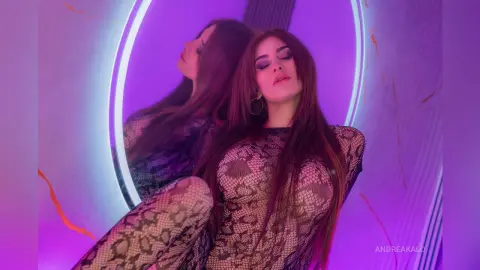 AndreaKalo poses in a black lace bodysuit against a purple glowing ring light, showing her long dark hair and cleavage.