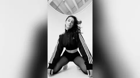 Stylish woman in black Adidas tracksuit posing confidently in a minimalist white studio setting.
