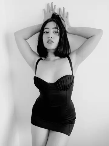 AnnaKomatsuu poses against a white wall wearing a black strapless mini dress with a sweetheart neckline, holding her hands up to her hair.