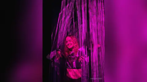 Smiling woman in a black crop top laughing under pink neon light through hanging string curtains.