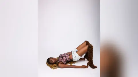 Fashionable woman in a pink patterned corset and denim shorts lying on a gold cushion against a white background.