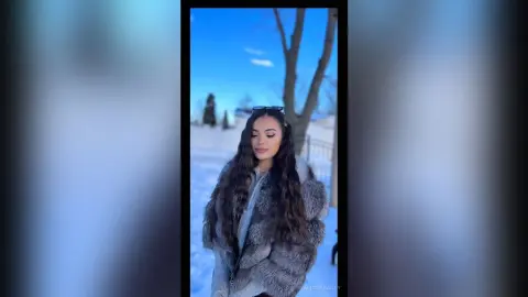 Young woman in a cozy grey faux fur coat posing in a bright snowy winter landscape under a blue sky.