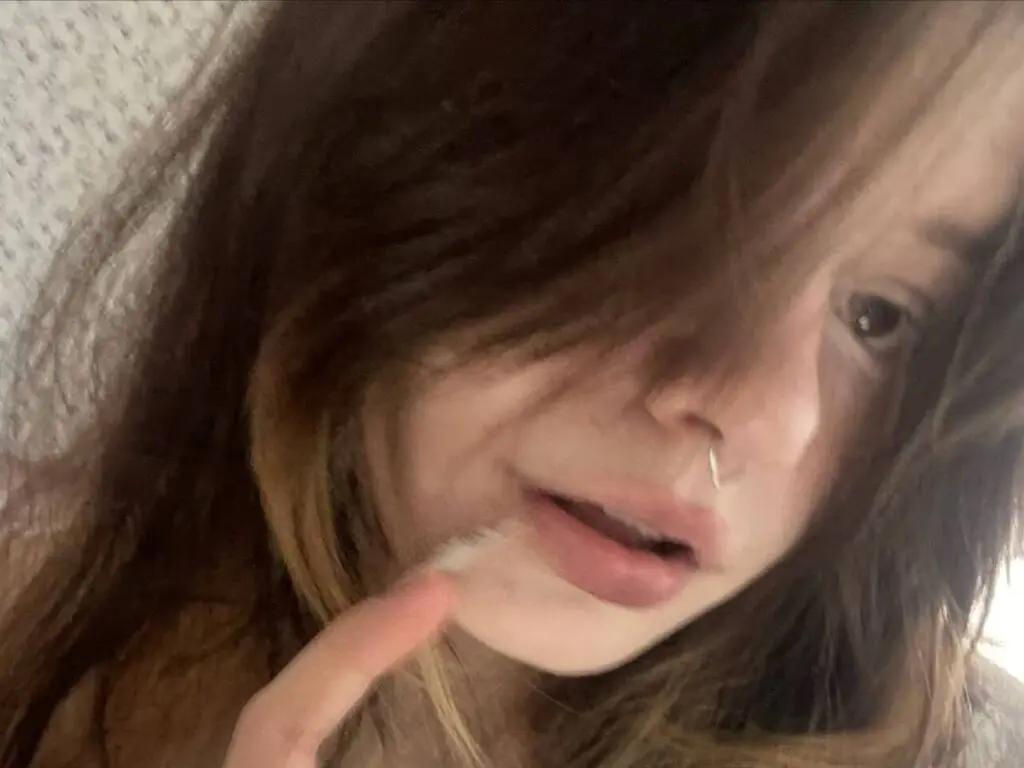 Close-up portrait of a young woman with long brown hair and a nose ring in a soft, moody indoor setting.