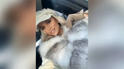 Smiling blonde woman in a cozy grey faux fur coat and cream newsboy cap sitting in a car.