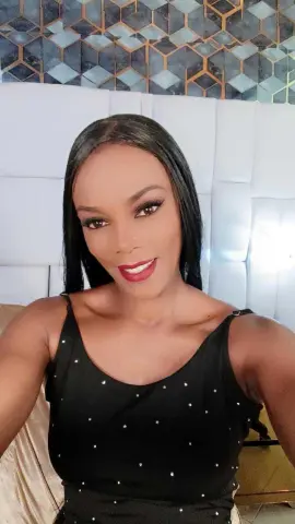 Smiling woman in a black tank top sitting against a white tufted headboard in a modern bedroom.