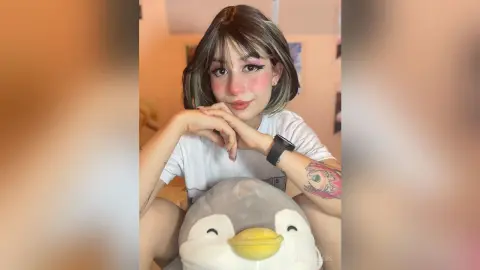 Smiling young woman with short brown hair in a white t-shirt sitting indoors with a cute penguin plushie.