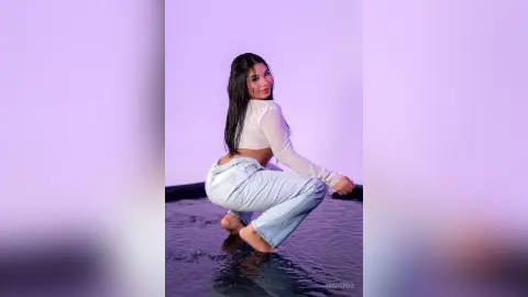 Smiling young woman in a white long-sleeve crop top and light blue jeans posing in a dreamy purple studio.