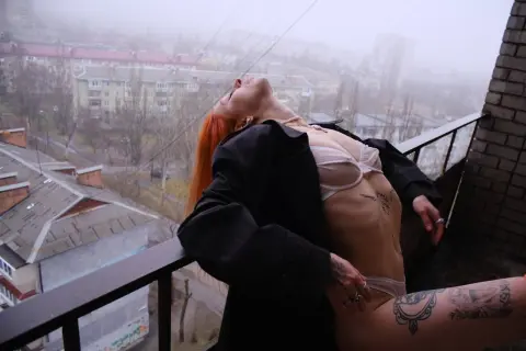 AlisaMoss leans back on a balcony in a black coat and white bra, showing her red hair and tattoos against a foggy city view.