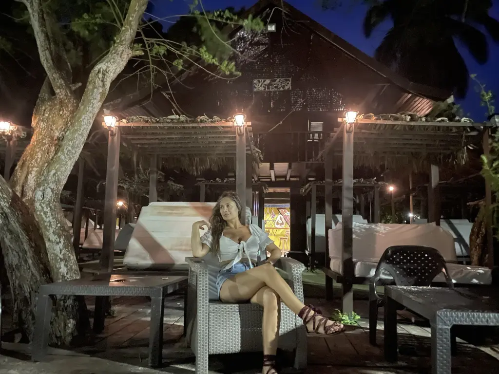 DaliaQuinn wearing a white top and denim shorts, sitting on a white lounge chair at a sandy beach setting.