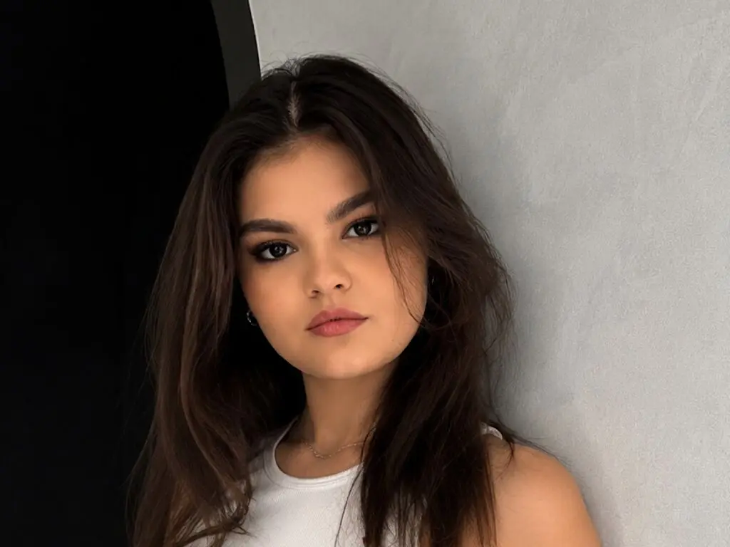 Young woman with long brown hair wearing a white tank top posing against a neutral grey background.