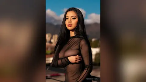 VickyPortman poses with long black hair wearing a sheer black long-sleeve bodysuit against a blue sky and mountain background.