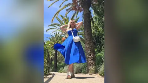 Blonde woman in a flowing royal blue maxi dress and white crossbody bag standing by palm trees in sunny weather.