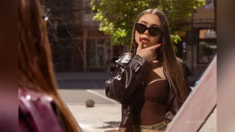 Stylish woman in a brown leather jacket and sunglasses reflecting in a mirror on a sunny city street.