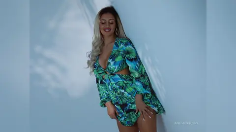 Anita poses against a light blue wall wearing a long-sleeved, low-cut green tropical print dress with a cut-out detail at the waist.