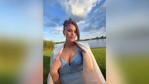 Elegant woman in a blue satin dress and white blazer posing outdoors by a lake under a blue sky.