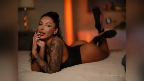 Tattooed woman in a black bodysuit lounging on a bed in warm, moody bedroom lighting.