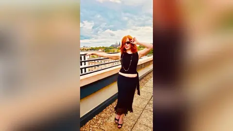 Redheaded woman in a chic black crop top and maxi skirt posing on a sunny urban balcony.