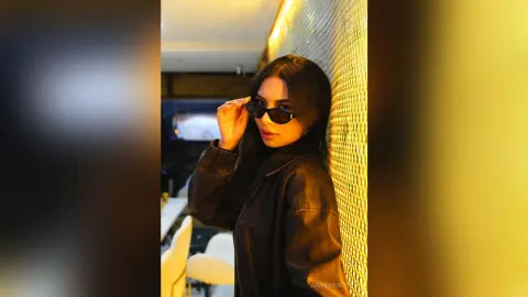 Stylish woman in a black leather jacket and sunglasses posing against a glowing gold textured wall.