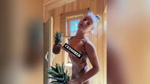 Young woman in a leopard print bikini holding pineapples in a rustic wooden cabin setting.