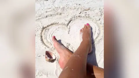 SinnaRay relaxes on the white sand with her bare feet resting inside a heart shape drawn in the sand.