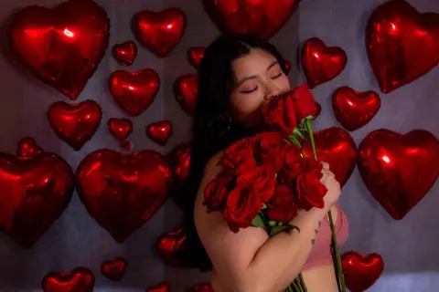 Young woman in pink top holding red roses against a backdrop of red heart balloons for a romantic Valentine's mood.
