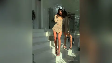 AnaMathers stands on white stairs wearing a tan button-down mini dress and black strappy heels.