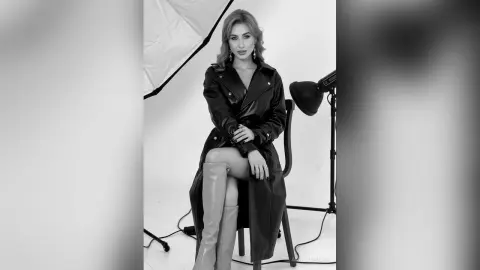 Stylish woman in a black leather trench coat and tall boots posing in a professional photo studio.