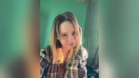 Smiling young woman with long blonde hair wearing a plaid flannel shirt in a cozy indoor setting.
