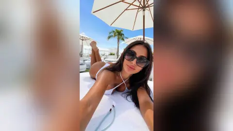 BiankaLeoness lies on a white lounge chair under a beach umbrella wearing a white bikini and sunglasses.