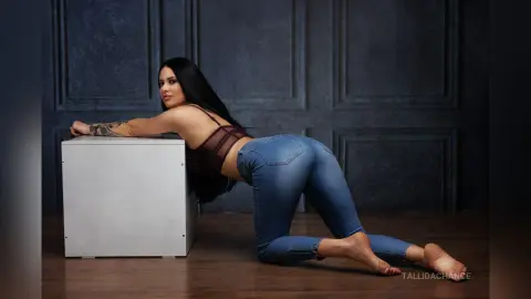 Woman in denim jeans and burgundy lace bodysuit posing on a white pedestal against a dark blue wall.