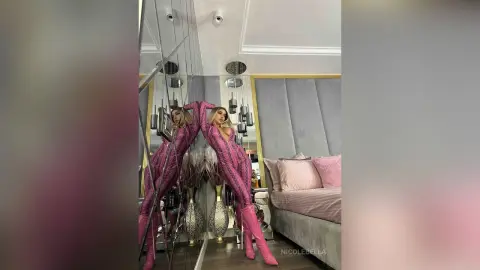 NicoleBella poses in a pink patterned bodysuit and matching pink boots against a mirrored wall in a luxury room.