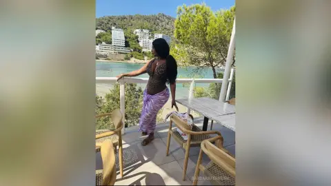 Woman in a patterned top and purple skirt relaxing on a sunny balcony overlooking a scenic coastal town.