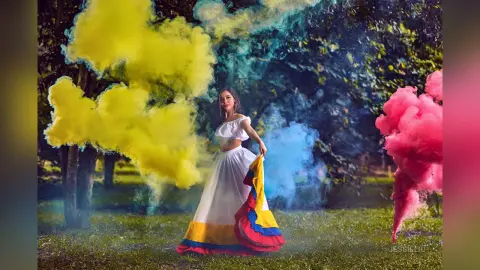 JessieLiu stands in a grassy field surrounded by thick clouds of yellow, blue, and pink smoke, wearing a white off-the-shoulder crop top and a long white skirt with a yellow and red trim.