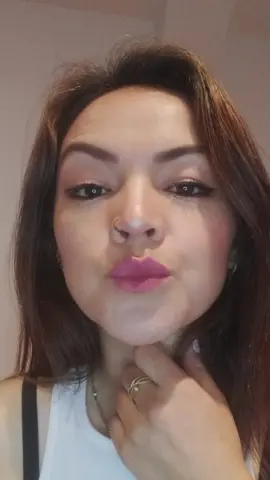 Woman with winged eyeliner and pink lipstick wearing a white tank top in a soft indoor setting.
