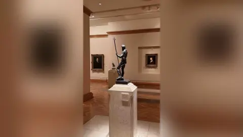 Bronze classical male statue standing on a marble pedestal in a quiet, modern art museum gallery.