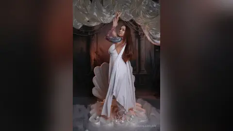 EsmeraldaClark poses in a white cut-out dress with her arms raised above her head, standing on a white shell prop surrounded by pearls.