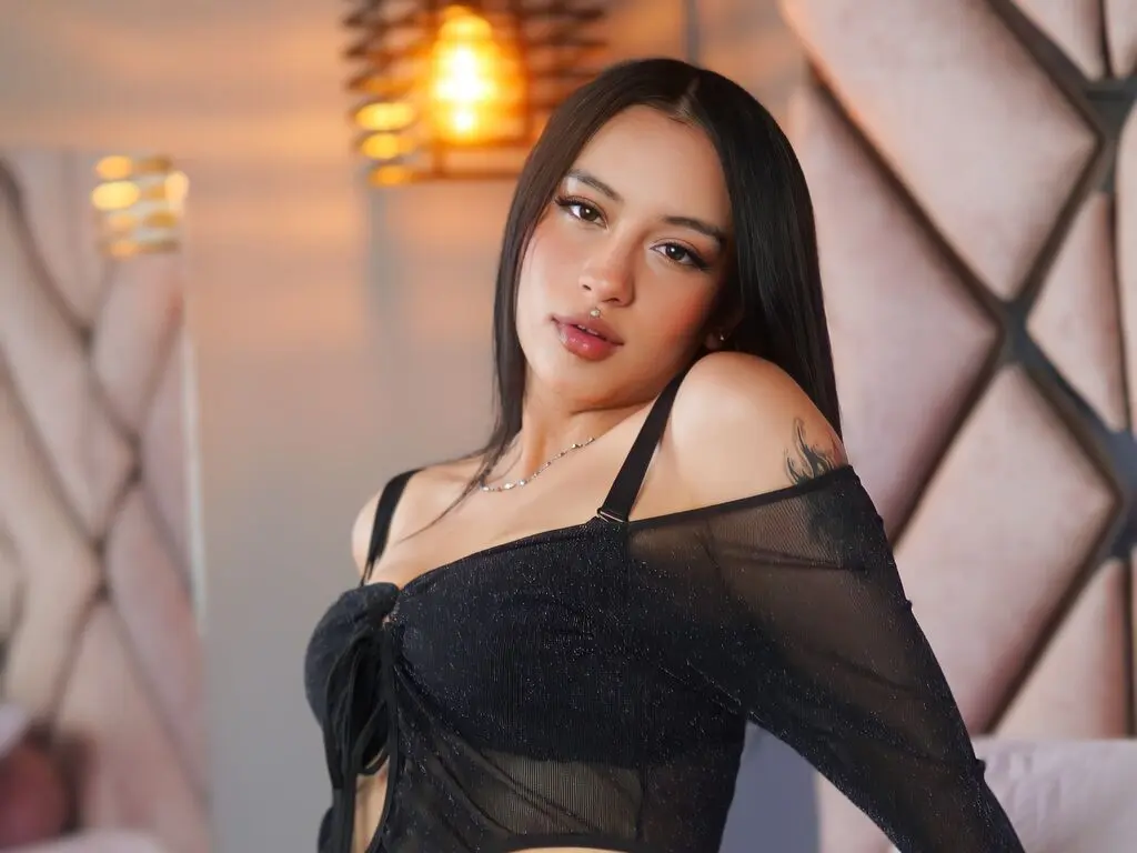 Young woman in a black sheer off-the-shoulder top posing with a soft, moody expression in a warm lit room.